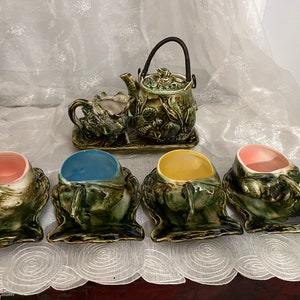 May include: A green and brown ceramic tea set with a teapot, creamer, and four cups. The cups are decorated with a frog design and have matching saucers.