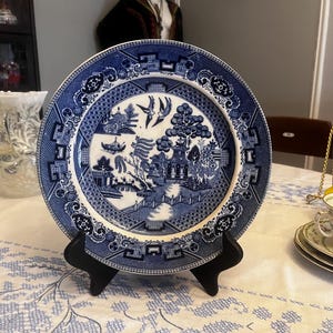 May include: A blue and white ceramic plate with a Willow pattern design, displayed on a black stand. The plate features a detailed landscape scene with pagodas, trees, and birds. The plate is set on a white tablecloth with a blue floral pattern.