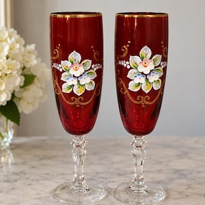 May include: A pair of tall, deep red glass champagne flutes featuring a hand-painted floral motif in white, green, and blue, accented with gold scrollwork and a gold rim. Each glass has a clear, faceted stem and a circular base for stability.