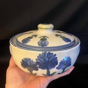 May include: A ceramic bowl with a lid, featuring a hand-painted floral design in shades of blue on a cream background. The bowl is round with a matching lid and a small knob. The floral pattern includes large flowers and smaller buds.