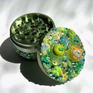 May include: A round, metallic green herb grinder with a multi-piece design. The lid is decorated with an assortment of green and yellow beads, star-shaped charms, and two small, sculpted cat figures resting in circular, translucent yellow frames.