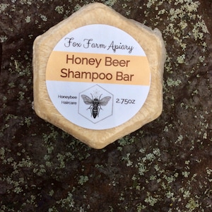 May include: A hexagonal Honey Beer Shampoo Bar from Fox Farm Apiary, wrapped in clear packaging. The label features the text "Honey Beer Shampoo Bar", "Honeybee Haircare", a bee illustration, and the weight, 2.75 oz.