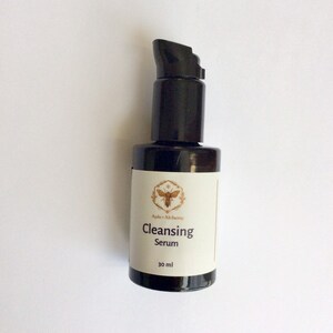 May include: A black glass bottle with a black pump top and a white label. The label says "Apis + Alchemy Cleansing Serum 30 ml".
