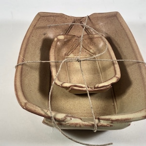 Puede incluir: Un juego de dos cuencos de cerámica, uno anidado dentro del otro, atados con una cuerda. Los cuencos son de color marrón claro con un esmalte moteado.