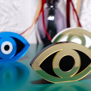 Op de afbeelding: Drie decoratieve boze oogcharms in blauw, goud en groen. De charmes zijn gevormd als een oog met een witte pupil en een zwarte iris.