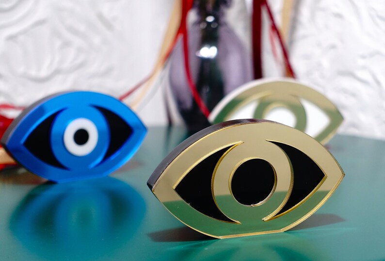 Op de afbeelding: Drie decoratieve boze oogcharms in blauw, goud en groen. De charmes zijn gevormd als een oog met een witte pupil en een zwarte iris.