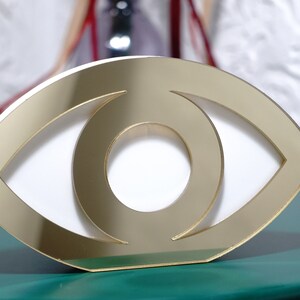 Op de afbeelding: Gouden oogvormig decoratief object met een witte binnenkant. Het object is gemaakt van gespiegeld acryl en heeft een modern ontwerp.