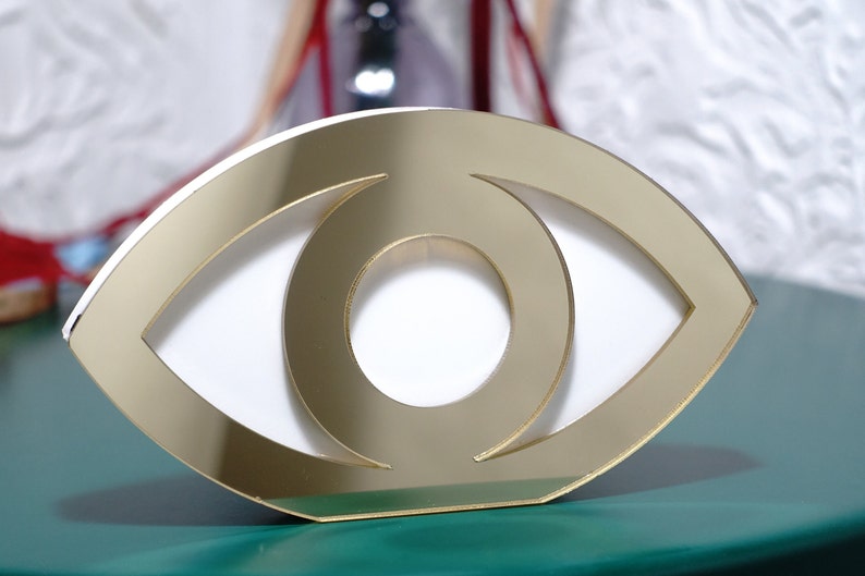 Op de afbeelding: Gouden oogvormig decoratief object met een witte binnenkant. Het object is gemaakt van gespiegeld acryl en heeft een modern ontwerp.