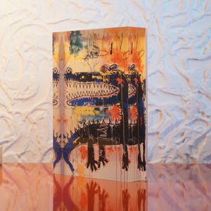 Puede incluir: Un bloque acr&iacute;lico colorido con una pintura de dos cocodrilos negros con la boca abierta y dientes afilados. El fondo es una mezcla de pintura amarilla, naranja, azul y blanca.