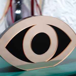 Op de afbeelding: Een goudkleurig, oogvormig decoratief object met een zwarte binnenkant. Het object is gemaakt van een glad, reflecterend materiaal.