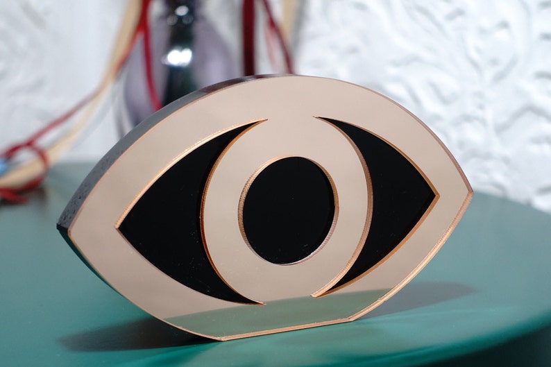 Op de afbeelding: Een goudkleurig, oogvormig decoratief object met een zwarte binnenkant. Het object is gemaakt van een glad, reflecterend materiaal.