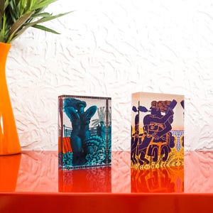 May include: Two decorative glass blocks with colorful illustrations sit on a red surface. One block features a blue figure, the other a purple and gold design. An orange vase with green foliage is to the left.