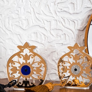 Puede incluir: Dos adornos decorativos con forma de granada hechos de material reflectante dorado con intrincados patrones recortados. Uno presenta una cuenta de ojo turco azul en el centro y el otro una cuenta de ojo turco con purpurina plateada, ambos con borla.