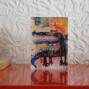 Puede incluir: Una pintura abstracta colorida de un cocodrilo con una boca azul y un cuerpo negro. El fondo es una mezcla de amarillo, naranja, rojo y azul.