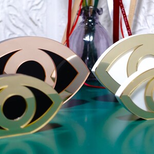 Op de afbeelding: Drie decoratieve oogvormige sculpturen gemaakt van gespiegeld acryl. De sculpturen zijn in verschillende maten en kleuren, waaronder goud, wit en ros&eacute; goud.
