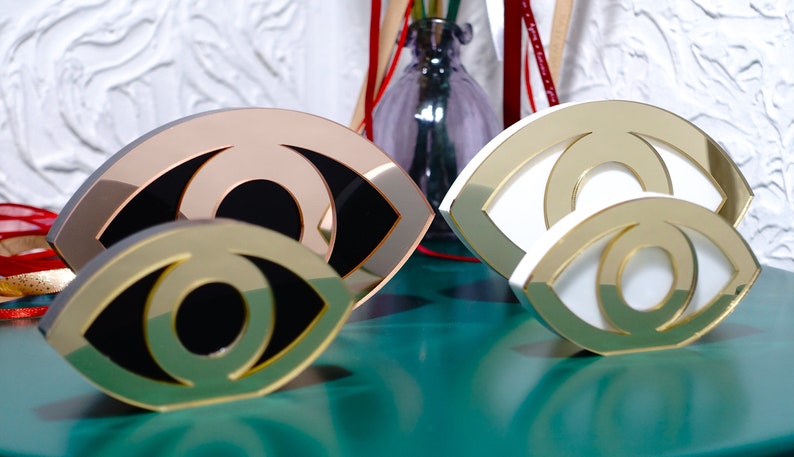 Op de afbeelding: Drie decoratieve oogvormige sculpturen gemaakt van gespiegeld acryl. De sculpturen zijn in verschillende maten en kleuren, waaronder goud, wit en ros&eacute; goud.