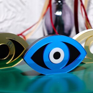 Op de afbeelding: Drie decoratieve boze oog-sculpturen gemaakt van gespiegeld acryl. De ogen zijn blauw, goud en wit met zwarte pupillen. De sculpturen zijn in een rij op een groen oppervlak gerangschikt.