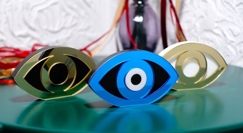Op de afbeelding: Drie decoratieve boze oog-sculpturen gemaakt van gespiegeld acryl. De ogen zijn blauw, goud en wit met zwarte pupillen. De sculpturen zijn in een rij op een groen oppervlak gerangschikt.