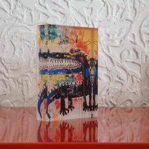 Puede incluir: Una pintura acr&iacute;lica abstracta colorida sobre un bloque de acr&iacute;lico transparente. La pintura presenta un caim&aacute;n negro y azul con una gran boca abierta y dientes blancos. El fondo es una mezcla de amarillo, naranja, azul y verde.