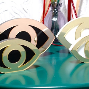 Op de afbeelding: Twee gouden oogvormige decoratieve sculpturen. Een sculptuur is wit met een gouden omtrek, de andere is zwart met een gouden omtrek. Beide sculpturen staan op een groen oppervlak.