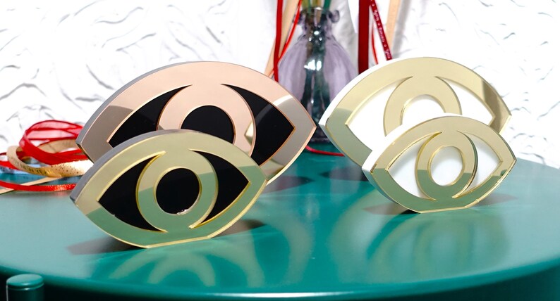 Op de afbeelding: Twee gouden oogvormige decoratieve sculpturen. Een sculptuur is wit met een gouden omtrek, de andere is zwart met een gouden omtrek. Beide sculpturen staan op een groen oppervlak.