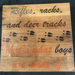 May include: Wooden sign with the text "Rifles, racks, and deer tracks that's what boys are made of" in various colors. The sign has a rustic look and features deer tracks painted on it.