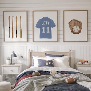 May include: Three framed baseball-themed prints hang above a bed. One print shows baseball bats, another a blue jersey with "JETT 11", and the third a baseball glove with a ball. The bed has a navy blue comforter and decorative pillows.