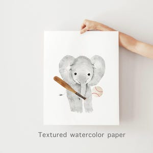 Może przedstawiać: Aquarela przedstawiająca szarego słonia trzymającego drewniany kij bejsbolowy i piłkę bejsbolową. Słoń stoi na białym tle.