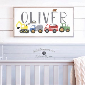 K&ouml;nnte beinhalten: Ein wei&szlig;es Holzschild mit dem Namen "Oliver" in grauen Buchstaben. Das Schild ist mit einer Reihe von bunten Spielzeugfahrzeugen verziert, darunter ein gelber Bagger, ein blauer LKW, ein grauer Betonmischer, ein rotes Feuerwehrauto und ein gr&uuml;ner Traktor. Das Schild h&auml;ngt an einer wei&szlig;en Holzwand &uuml;ber einem grauen Kinderbett.