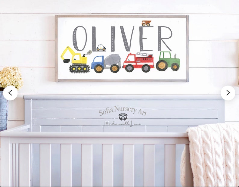 K&ouml;nnte beinhalten: Ein wei&szlig;es Holzschild mit dem Namen "Oliver" in grauen Buchstaben. Das Schild ist mit einer Reihe von bunten Spielzeugfahrzeugen verziert, darunter ein gelber Bagger, ein blauer LKW, ein grauer Betonmischer, ein rotes Feuerwehrauto und ein gr&uuml;ner Traktor. Das Schild h&auml;ngt an einer wei&szlig;en Holzwand &uuml;ber einem grauen Kinderbett.