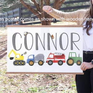 K&ouml;nnte beinhalten: Ein wei&szlig;er Leinwand-Poster mit dem Namen "CONNOR" in Grau auf einer grauen Stra&szlig;e gedruckt. Der Poster zeigt einen gelben Bagger, einen blauen Kipplaster, einen grauen Betonmischer, ein rotes Feuerwehrauto und einen gr&uuml;nen Traktor. Der Poster h&auml;ngt an einer Holzleiste.