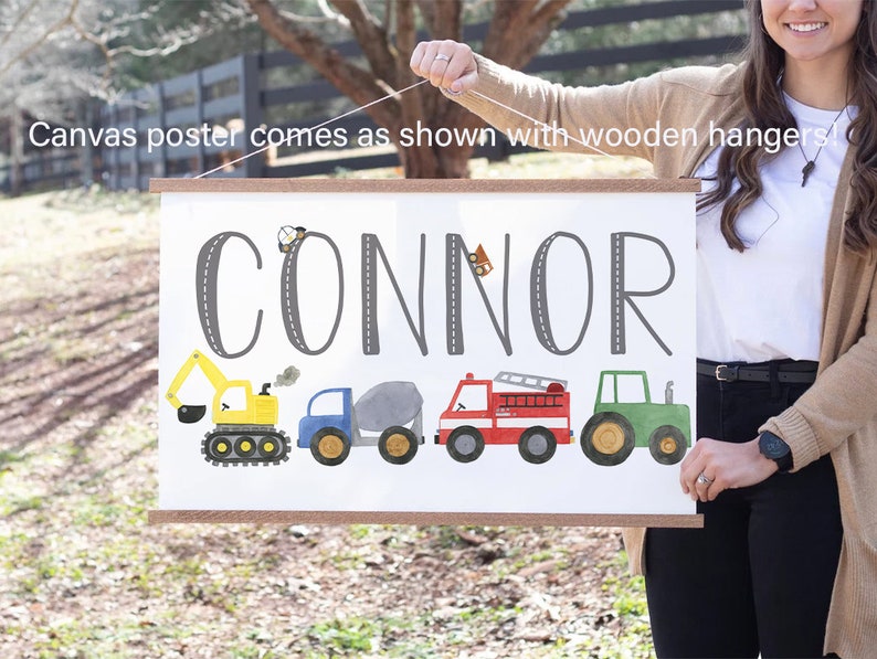 K&ouml;nnte beinhalten: Ein wei&szlig;er Leinwand-Poster mit dem Namen "CONNOR" in Grau auf einer grauen Stra&szlig;e gedruckt. Der Poster zeigt einen gelben Bagger, einen blauen Kipplaster, einen grauen Betonmischer, ein rotes Feuerwehrauto und einen gr&uuml;nen Traktor. Der Poster h&auml;ngt an einer Holzleiste.