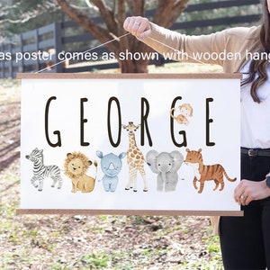 May include: A white canvas poster with the name "GEORGE" printed in black. The poster features a line of cartoon animals including a zebra, lion, rhinoceros, giraffe, elephant, and tiger. The poster is hanging from a wooden dowel rod.