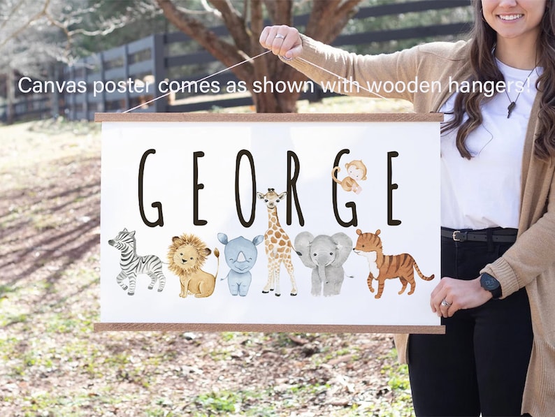 May include: A white canvas poster with the name "GEORGE" printed in black. The poster features a line of cartoon animals including a zebra, lion, rhinoceros, giraffe, elephant, and tiger. The poster is hanging from a wooden dowel rod.