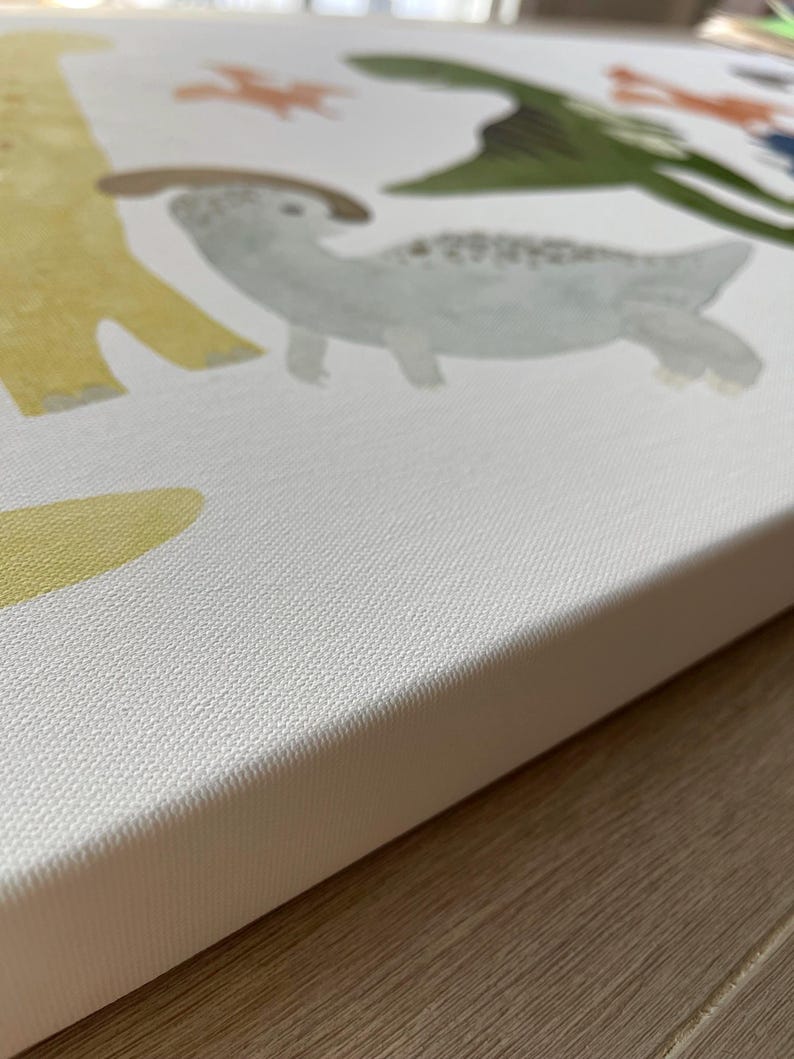 Pode incluir: Um close-up da borda de uma tela branca com uma superf&iacute;cie texturizada. A tela tem uma pintura de um dinossauro amarelo, um dinossauro verde e um dinossauro cinza com manchas marrons.