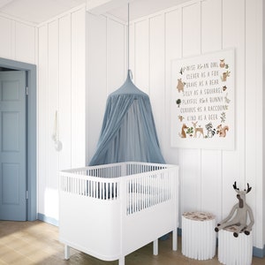 May include: A white crib with a blue canopy hanging above it. A framed print with the text "Wise as an owl, Clever as a fox, Brave as a bear, Silly as a squirrel, Playful as a bunny, Curious as a raccoon, Kind as a deer" is on the wall. There are two wooden stools with a stuffed deer toy sitting on one.