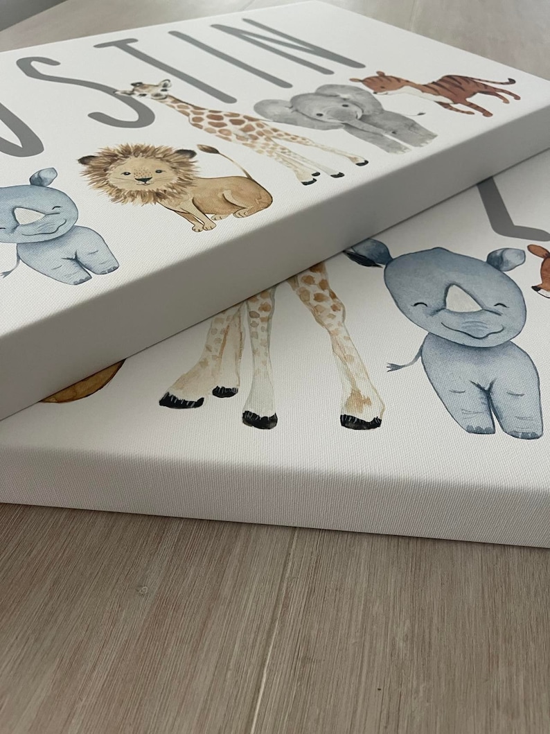 May include: A white canvas with the name "Justin" printed in gray letters. The canvas features a line of watercolor animal illustrations, including a rhinoceros, a lion, a giraffe, an elephant, and a tiger.