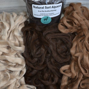 May include: Three piles of natural Suri alpaca roving in beige, brown, and light brown. The bag in the center reads "Natural Suri Alpaca 4 oz Pin Drafted Roving Celebrate your Creativity! Specialty fiber for spinning, felting, dyeing & more."