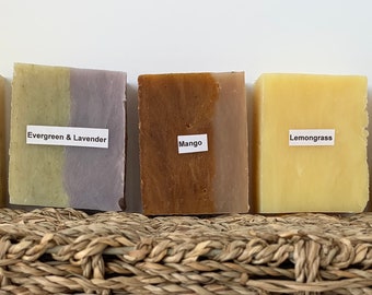 May include: Five bars of handmade soap with different scents: Lavender, Evergreen & Lavender, Mango, Lemongrass, and Mountain Air. The bars are arranged in a row on a woven basket.