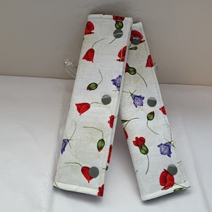 Op de afbeelding: Twee witte stoffen gordelhoezen met een bloemenpatroon van rode klaprozen, paarse klokjes en groene bladeren. De hoezen hebben grijze knopen.