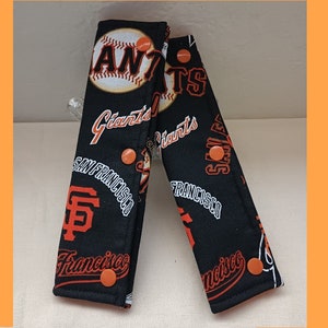 Puede incluir: Dos correas de tela negra con botones naranjas y un patrón repetido del logotipo de los Gigantes de San Francisco, pelotas de béisbol y las palabras "Giants", "San Francisco" y "Francisco".