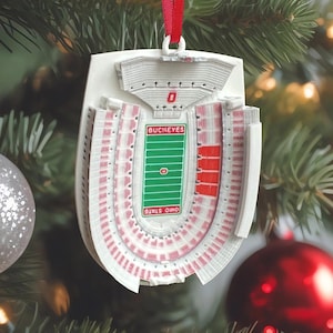 May include: A white, three-dimensional stadium ornament featuring a green football field with red and white details. The field displays the text BUCKEYES and OHIO STATE. The structure includes red seating sections and is suspended by a red ribbon.