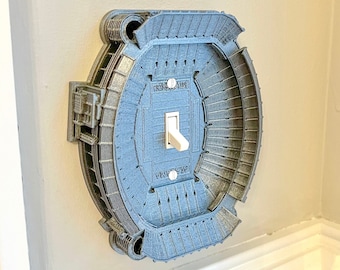 May include: A light switch cover shaped like a stadium with a white toggle switch. The switch cover is gray and has a detailed design of the stadium.