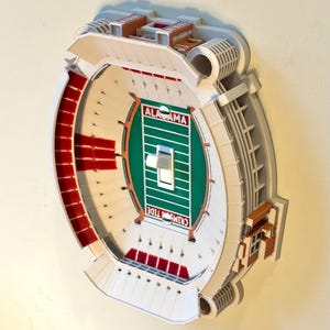 Może przedstawiać: Trójwymiarowa osłona włącznika światła w kształcie stadionu futbolowego. W centrum znajduje się zielone boisko z białymi liniami, napis ALABAMA i CRIMSON TIDE oraz biały przełącznik. Trybuny stadionu są białe z czerwonymi akcentami.