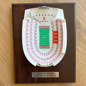 Może przedstawiać: Tr&oacute;jwymiarowy model stadionu Ohio, domu Ohio State Buckeyes, na drewnianej tabliczce. Model jest biały z czerwonymi siedzeniami i zielonym boiskiem. Tabliczka zawiera napis "Ohio Stadium" i "Ohio State Buckeyes".