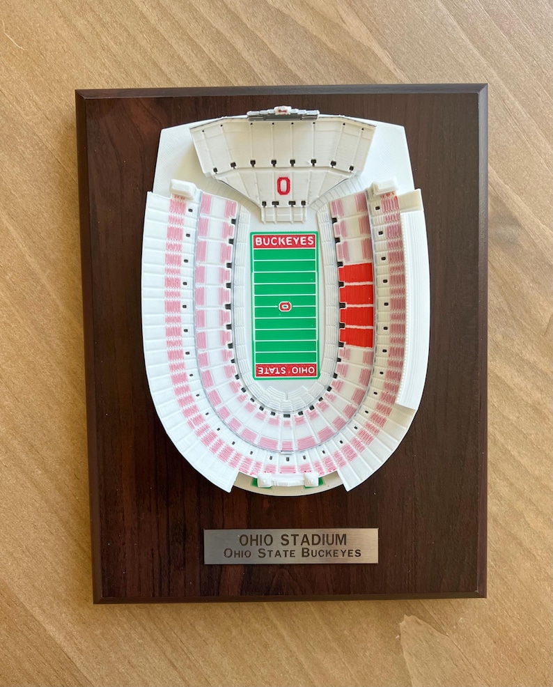 Może przedstawiać: Tr&oacute;jwymiarowy model stadionu Ohio, domu Ohio State Buckeyes, na drewnianej tabliczce. Model jest biały z czerwonymi siedzeniami i zielonym boiskiem. Tabliczka zawiera napis "Ohio Stadium" i "Ohio State Buckeyes".