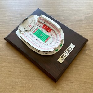 Może przedstawiać: Miniaturowa replika stadionu Ohio, domu Ohio State Buckeyes, na ciemnej drewnianej tabliczce. Stadion jest biały z czerwonymi i r&oacute;żowymi detalami. Tabliczka ma złotą etykietę z tekstem "Ohio Stadium Ohio State Buckeyes".