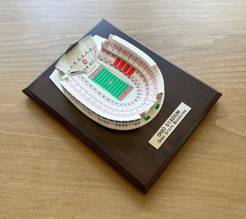 Może przedstawiać: Miniaturowa replika stadionu Ohio, domu Ohio State Buckeyes, na ciemnej drewnianej tabliczce. Stadion jest biały z czerwonymi i r&oacute;żowymi detalami. Tabliczka ma złotą etykietę z tekstem "Ohio Stadium Ohio State Buckeyes".