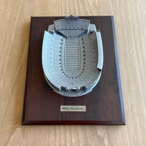 Może przedstawiać: Tr&oacute;jwymiarowy model stadionu Ohio Stadium, domu drużyny Ohio State Buckeyes, na ciemnej drewnianej tabliczce. Model jest szary i przedstawia miejsca siedzące na stadionie, boisko i tablicę wynik&oacute;w. Na tabliczce widnieje napis "Ohio Stadium".