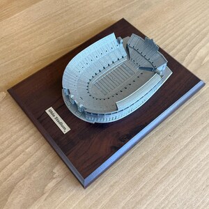 Może przedstawiać: Srebrny model stadionu Ohio na ciemnym drewnianym cokole. Na cokole znajduje się tabliczka z napisem "Ohio Stadium."